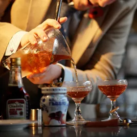 A bartender pouring a stirred cocktail from a mixing glass into two coupes
