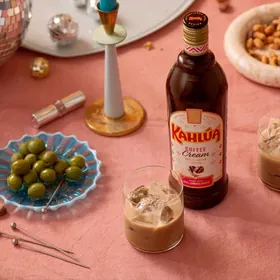 A bottle of Kahlúa Coffee Cream beside a glass of it over ice, with olives and party snacks