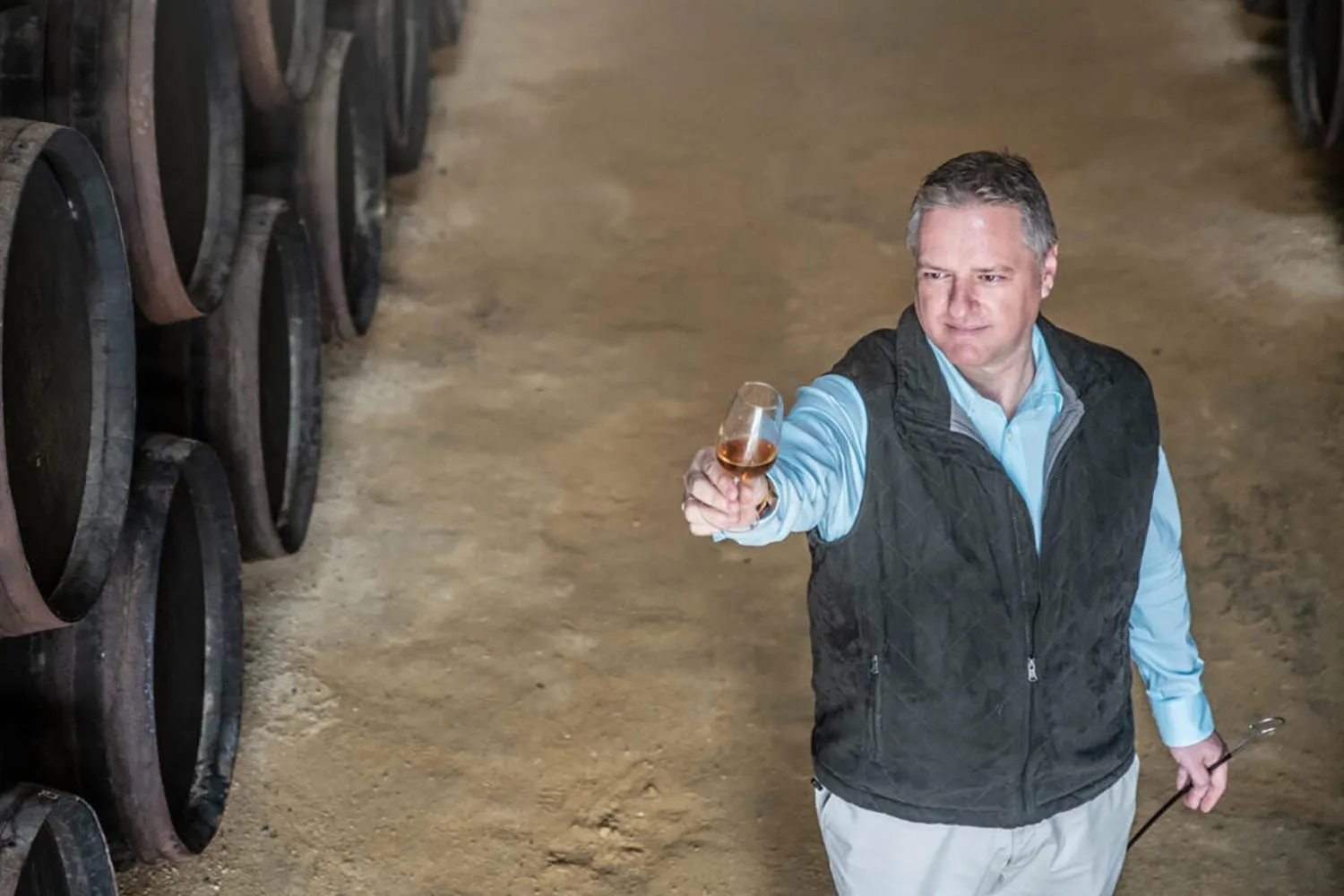 A maker holding a tasting glass of amber spirit up to the light in a barrel-lined cellar
